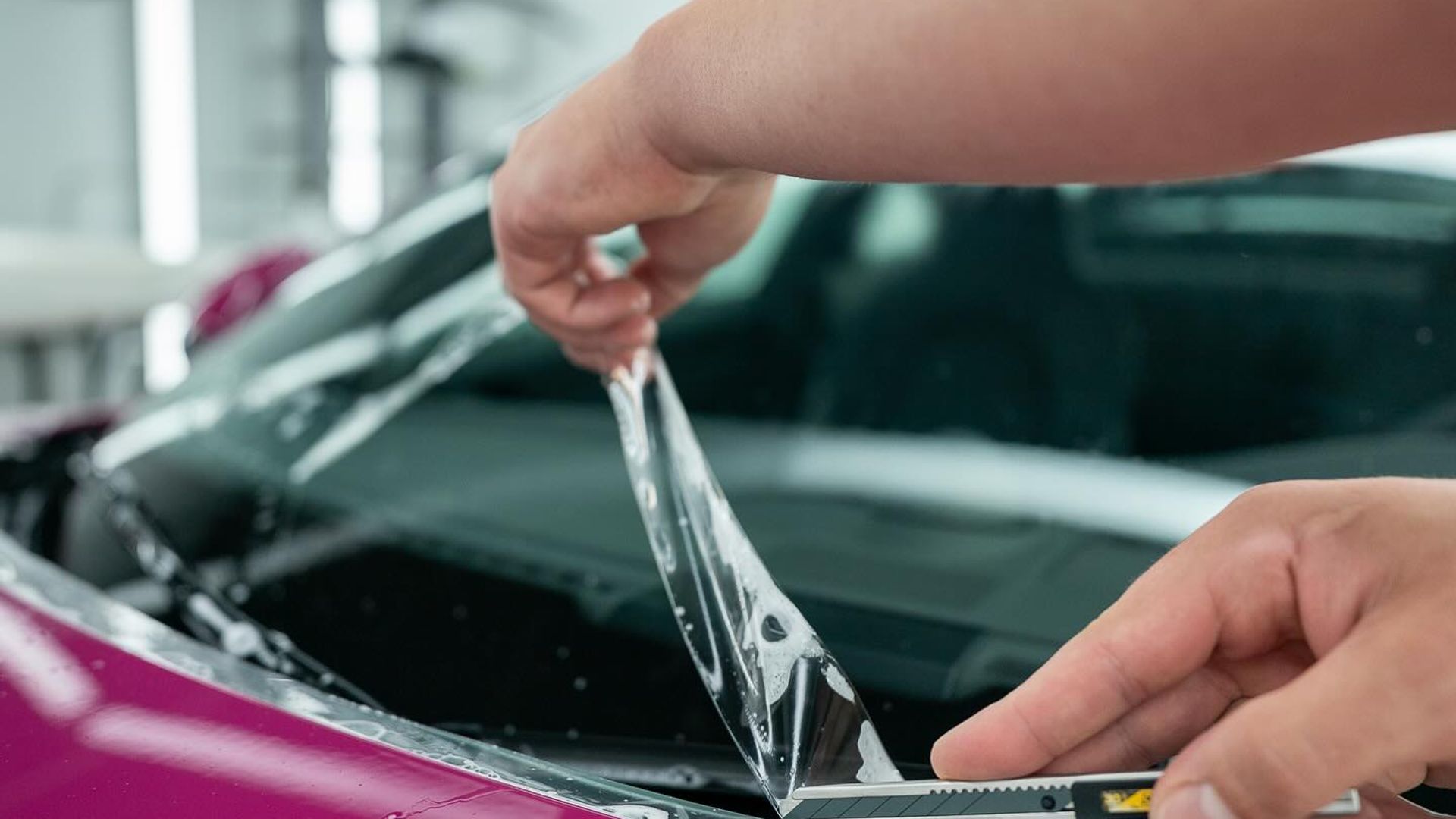 Hands precision-cutting PPF film on a pink car with a detail knife at EMWRAPS